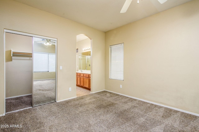 unfurnished bedroom with connected bathroom, light colored carpet, a closet, arched walkways, and a ceiling fan