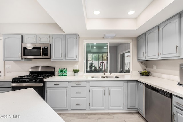 kitchen with a raised ceiling, appliances with stainless steel finishes, light countertops, gray cabinetry, and a sink