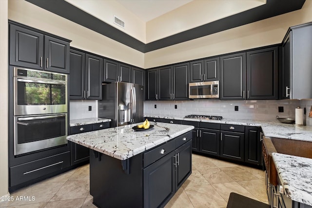 kitchen with stainless steel appliances, a kitchen island, light stone countertops, dark cabinetry, and backsplash