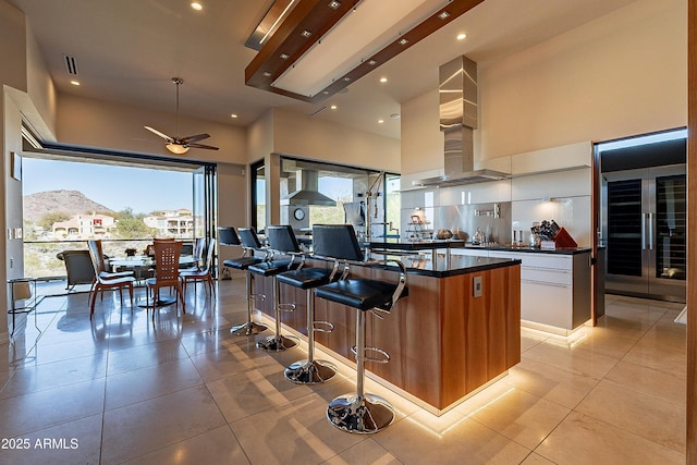 kitchen featuring wall chimney exhaust hood, light tile patterned floors, a mountain view, a kitchen island with sink, and white cabinets