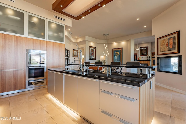 kitchen with light tile patterned floors, white cabinets, an island with sink, and stainless steel double oven