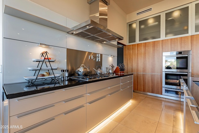 kitchen featuring appliances with stainless steel finishes, island range hood, light tile patterned floors, and dark stone countertops