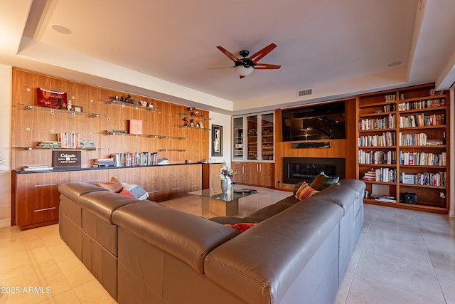 tiled living room featuring built in shelves, ceiling fan, wooden walls, and a raised ceiling