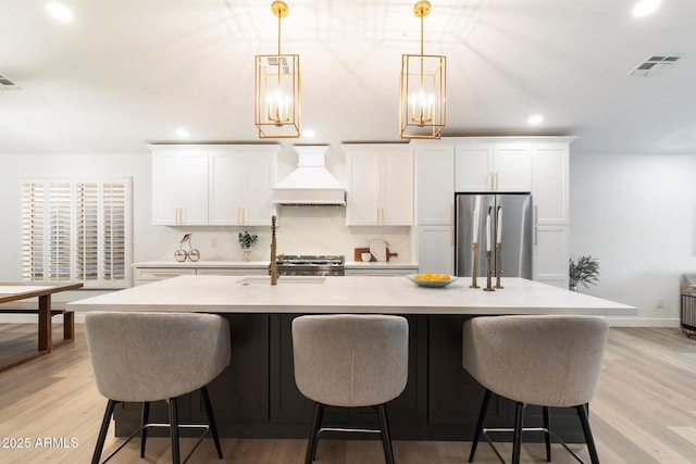 kitchen with stainless steel refrigerator, white cabinetry, custom exhaust hood, and a center island with sink