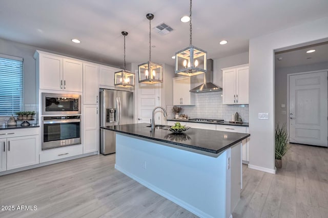 kitchen with wall chimney exhaust hood, decorative light fixtures, appliances with stainless steel finishes, a kitchen island with sink, and white cabinets