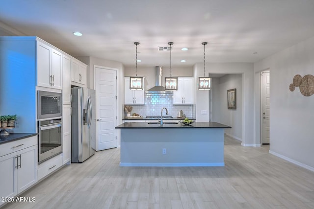 kitchen with hanging light fixtures, wall chimney range hood, stainless steel appliances, a kitchen island with sink, and white cabinets