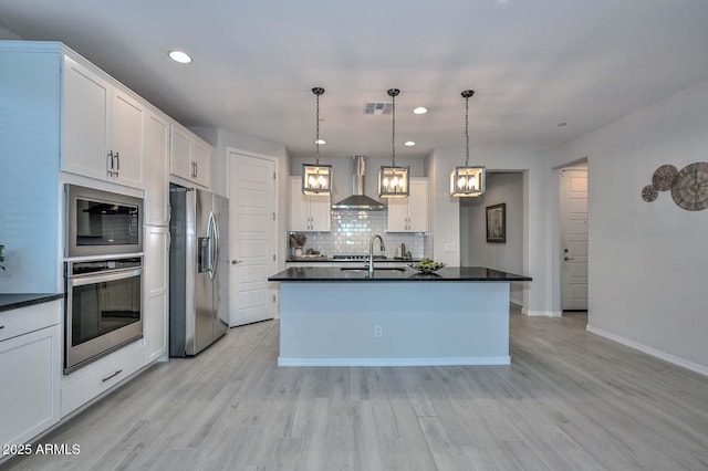 kitchen featuring a center island with sink, wall chimney range hood, pendant lighting, stainless steel appliances, and white cabinets