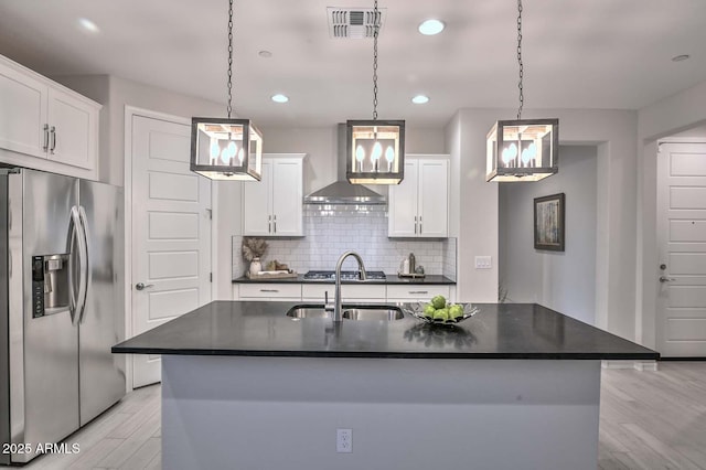 kitchen featuring appliances with stainless steel finishes, a kitchen island with sink, hanging light fixtures, white cabinetry, and wall chimney exhaust hood