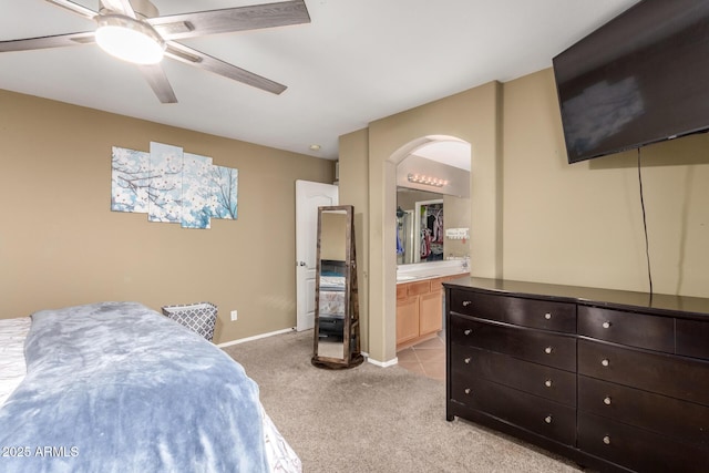 bedroom featuring ceiling fan, light colored carpet, arched walkways, baseboards, and ensuite bathroom