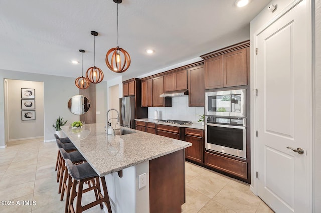 19908 W Montecito Ave, Litchfield Park, AZ 85340 - kitchen featuring appliances with stainless steel finishes, under cabinet range hood, light tile patterned flooring, a kitchen bar, and a center island with sink