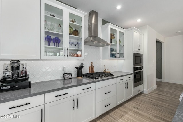 kitchen featuring appliances with stainless steel finishes, white cabinetry, backsplash, wall chimney exhaust hood, and light wood-type flooring