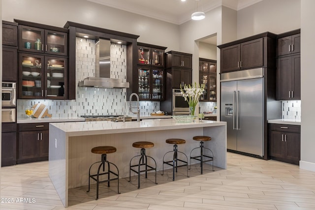 kitchen featuring wall chimney exhaust hood, stainless steel appliances, dark brown cabinets, light countertops, and backsplash