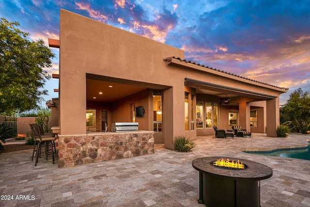 patio terrace at dusk featuring a fire pit, an outdoor kitchen, an outdoor bar, ceiling fan, and area for grilling