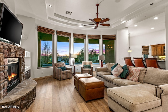 living room with a tray ceiling, a stone fireplace, ceiling fan, and light hardwood / wood-style floors