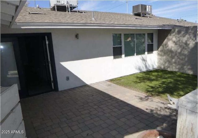 back of house featuring stucco siding, central AC, and roof with shingles