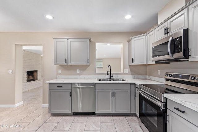 kitchen with gray cabinetry, light stone countertops, sink, stainless steel appliances, and a stone fireplace