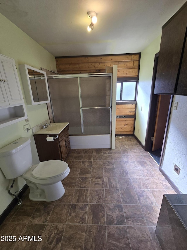 bathroom with bath / shower combo with glass door, toilet, stone finish floor, a sink, and baseboards