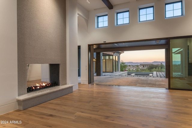 unfurnished living room with a warm lit fireplace, beamed ceiling, light wood-type flooring, and a towering ceiling