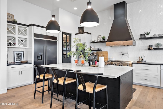 kitchen with wall chimney exhaust hood, hanging light fixtures, a center island with sink, white cabinetry, and stainless steel built in refrigerator