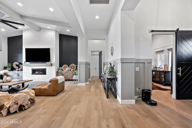 living room with beam ceiling, a large fireplace, a barn door, light wood-type flooring, and ceiling fan