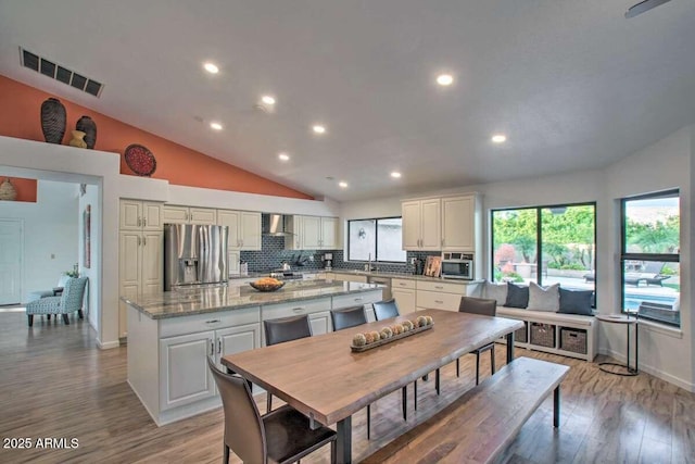 kitchen with stainless steel refrigerator with ice dispenser, wall chimney exhaust hood, tasteful backsplash, light stone counters, and a center island