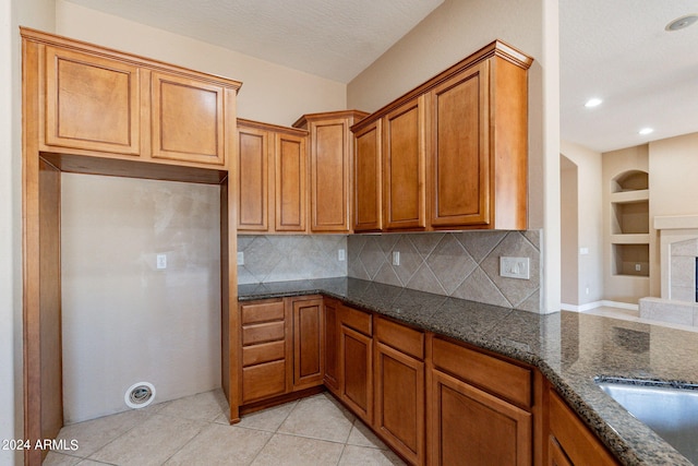 kitchen featuring tasteful backsplash, a fireplace with raised hearth, dark stone counters, light tile patterned floors, and brown cabinets