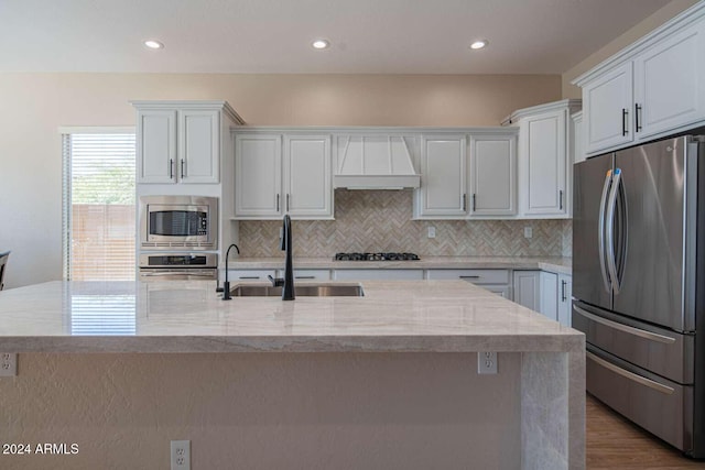 kitchen featuring premium range hood, white cabinets, a center island with sink, sink, and stainless steel appliances