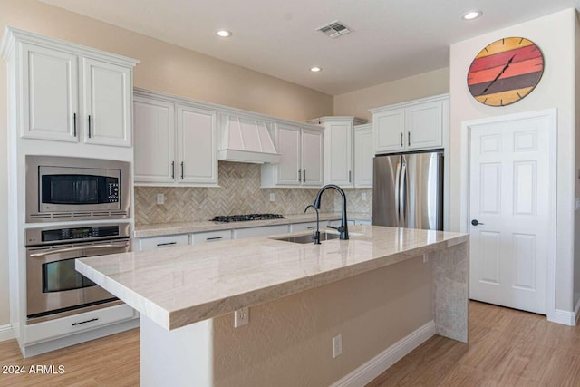 kitchen featuring sink, stainless steel appliances, premium range hood, a center island with sink, and white cabinets