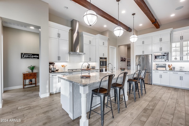 kitchen with pendant lighting, an island with sink, beam ceiling, wall chimney exhaust hood, and appliances with stainless steel finishes