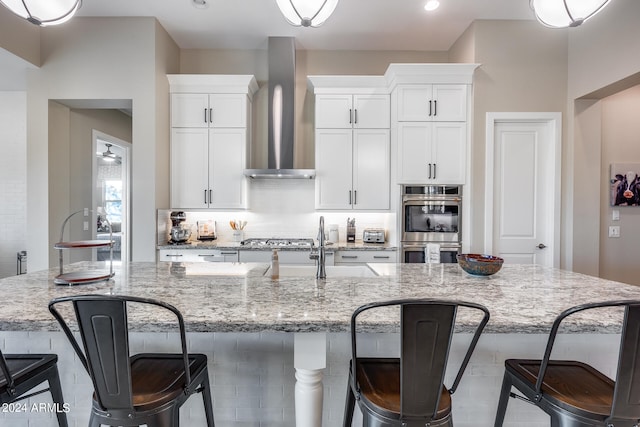 kitchen with a large island with sink, white cabinets, wall chimney exhaust hood, appliances with stainless steel finishes, and light stone countertops