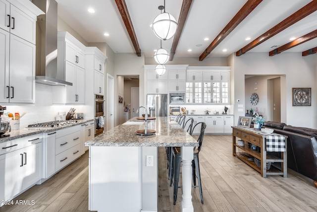 kitchen with light stone counters, beamed ceiling, an island with sink, wall chimney exhaust hood, and appliances with stainless steel finishes