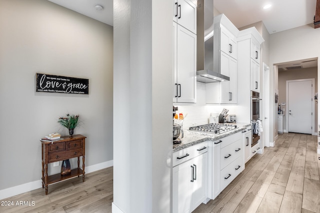 kitchen with light wood-type flooring, wall chimney exhaust hood, white cabinetry, stainless steel appliances, and light stone countertops