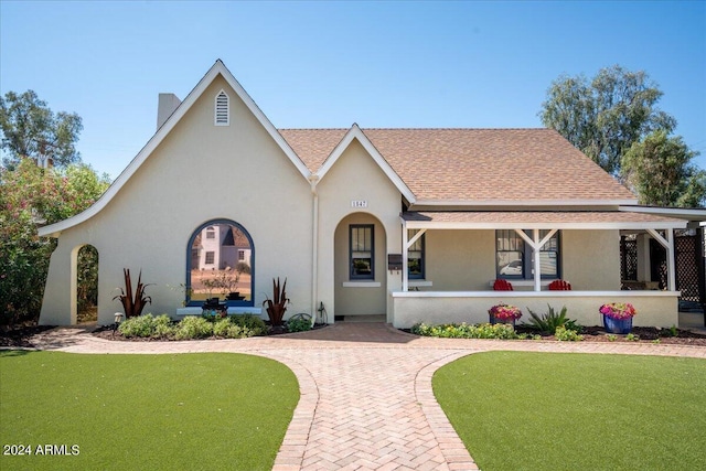 tudor house with a front yard, covered porch, roof with shingles, and stucco siding