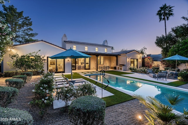 back of property at dusk with a tiled roof, a chimney, an outdoor pool, a patio area, and stucco siding