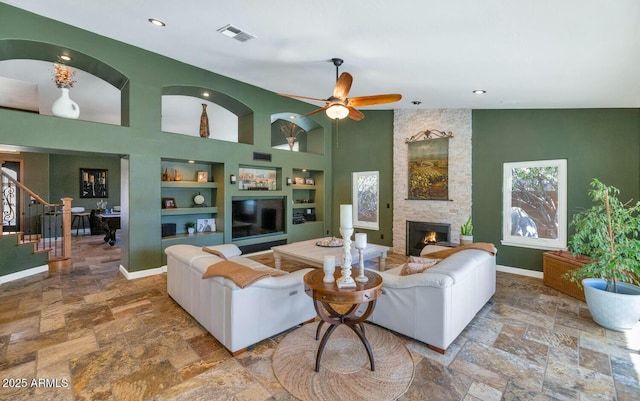 living room featuring ceiling fan, a high ceiling, built in features, and a stone fireplace