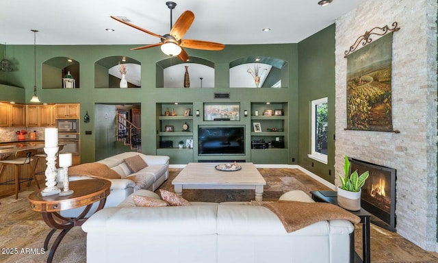 living room with ceiling fan, built in shelves, a fireplace, and a towering ceiling