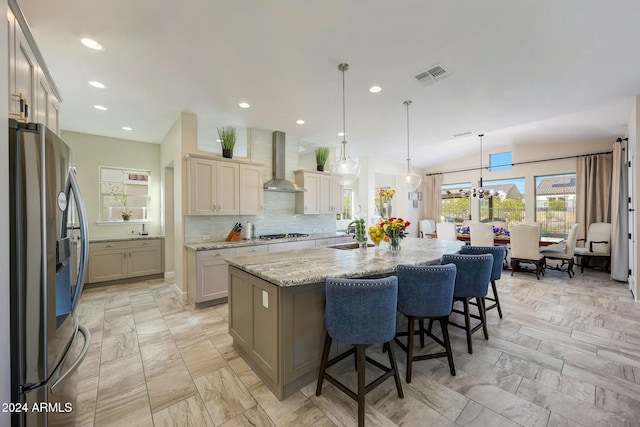 kitchen featuring light stone counters, wall chimney exhaust hood, stainless steel appliances, a kitchen island with sink, and hanging light fixtures