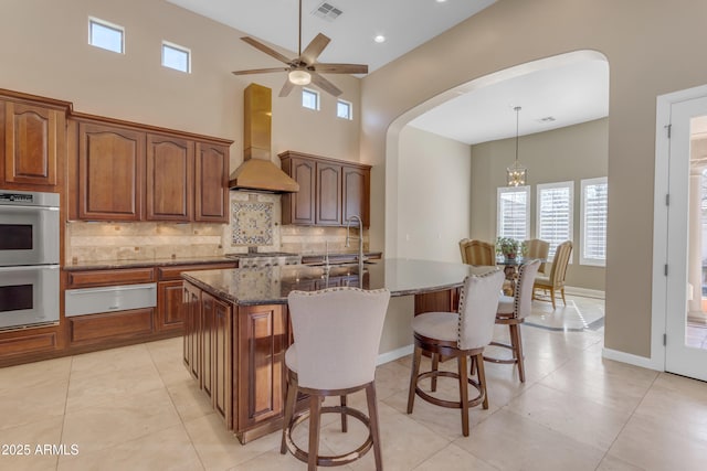 kitchen featuring hanging light fixtures, custom exhaust hood, tasteful backsplash, an island with sink, and stainless steel double oven