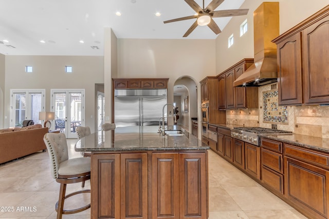 kitchen with appliances with stainless steel finishes, sink, wall chimney exhaust hood, a kitchen island with sink, and decorative backsplash