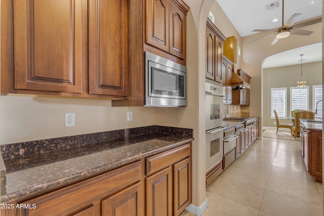 kitchen with ceiling fan with notable chandelier, wall chimney exhaust hood, stainless steel appliances, dark stone counters, and light tile patterned flooring