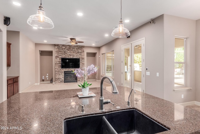 kitchen featuring dark stone countertops, hanging light fixtures, sink, a stone fireplace, and ceiling fan