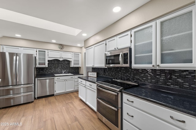 kitchen with a sink, light wood-style floors, appliances with stainless steel finishes, backsplash, and glass insert cabinets