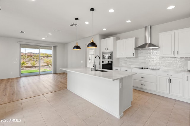 kitchen with sink, stainless steel microwave, an island with sink, wall oven, and wall chimney range hood