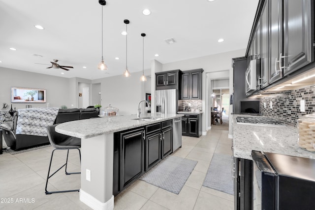 kitchen featuring light tile patterned floors, a kitchen island with sink, stainless steel appliances, light stone counters, and decorative light fixtures