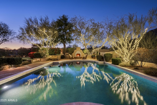 pool at dusk with an outdoor pool, a patio, a lit fireplace, and fence