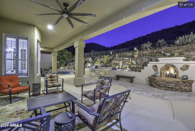 patio terrace at dusk with an outdoor stone fireplace, area for grilling, and ceiling fan