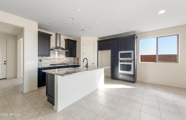 kitchen featuring light stone counters, dark cabinets, appliances with stainless steel finishes, wall chimney exhaust hood, and a center island with sink