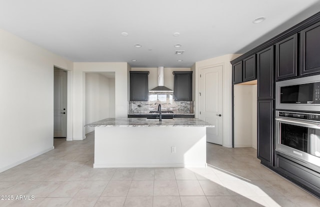 kitchen featuring built in microwave, oven, a center island with sink, and wall chimney range hood