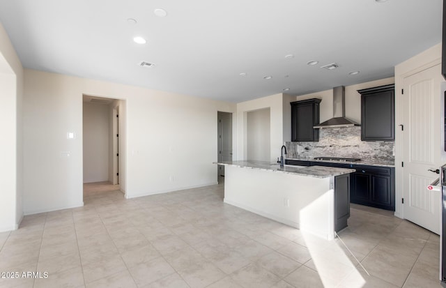 kitchen featuring a center island with sink, visible vents, wall chimney exhaust hood, light stone counters, and stainless steel gas cooktop