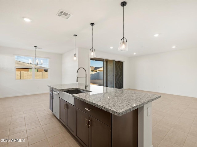 kitchen with sink, light stone counters, an island with sink, dark brown cabinets, and pendant lighting